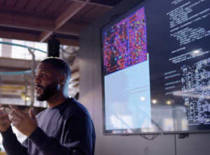 A man speaking in front of a TV screen.