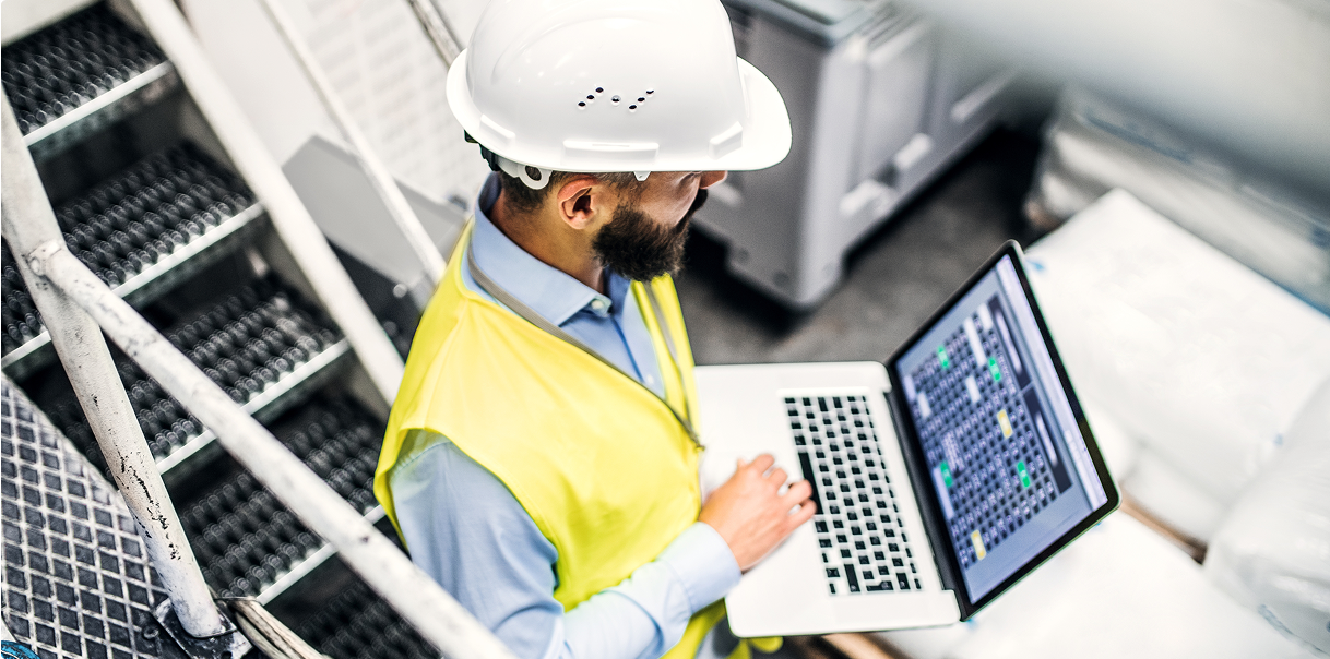A man wearing a construction hard hat walking down the staircase holding a laptop.