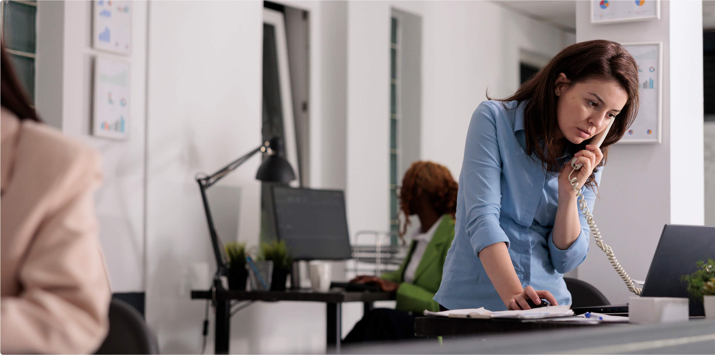 A woman using a landline phone and looking over a laptop screen.