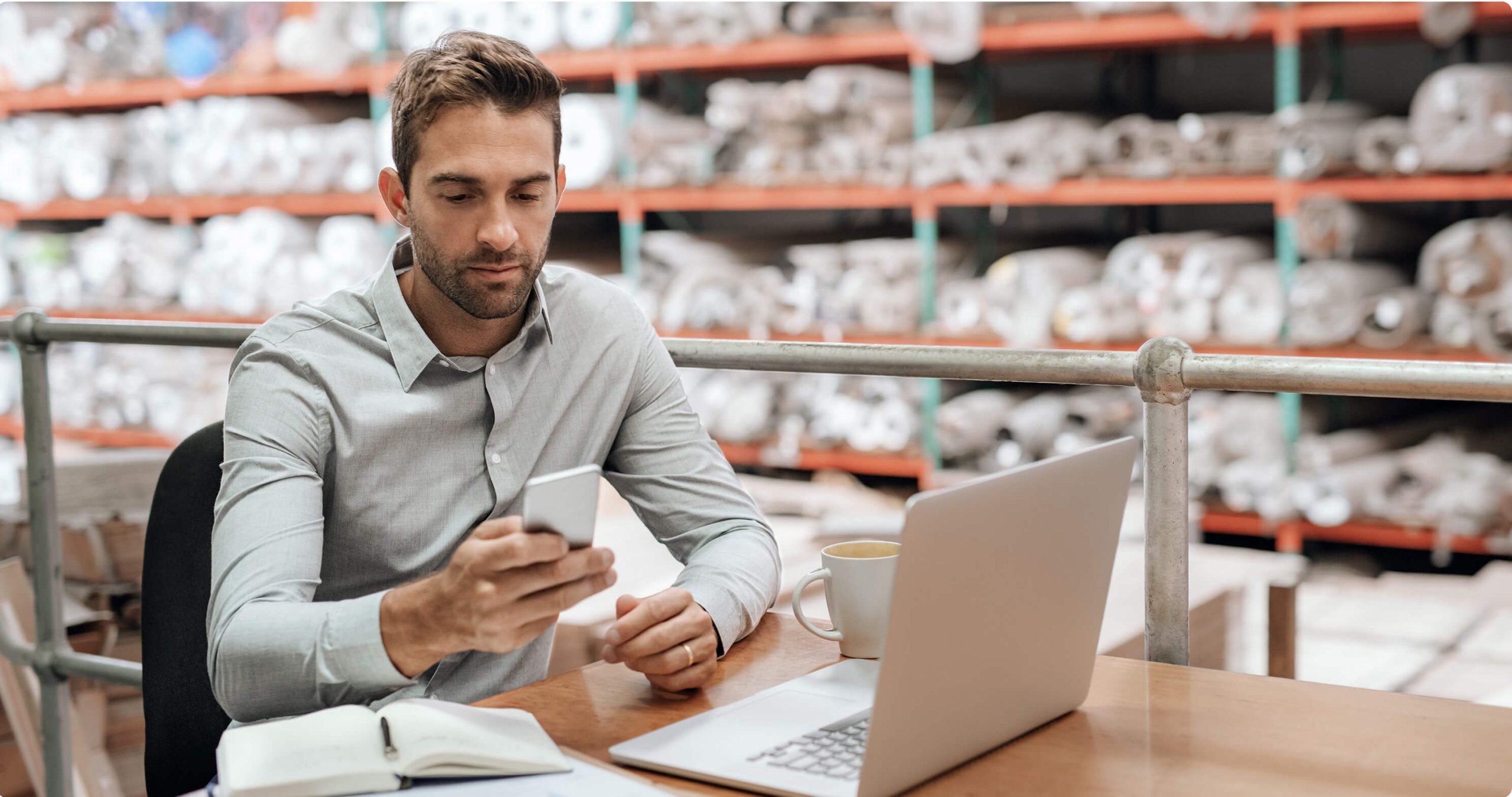 A man sitting at a desk in a warehouse environment, holding a phone in one hand, with a laptop on the desk.