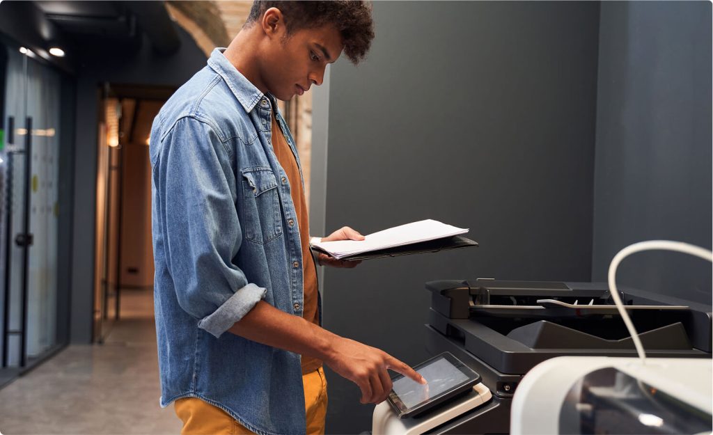 A person standing at a photocopier machine, pressing a touchscreen for operation.
