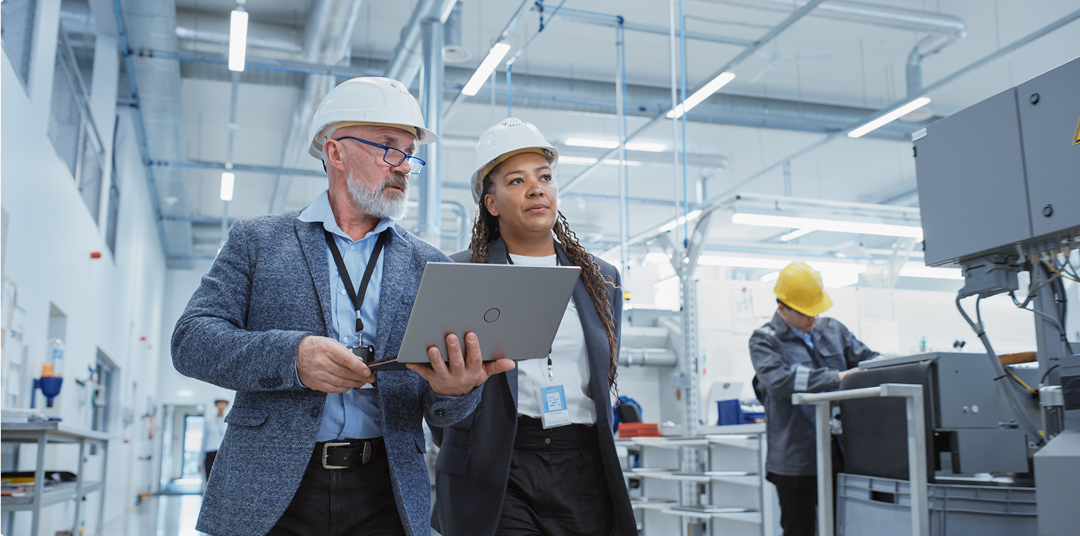 Two workers wearing hard hats, one carrying a laptop. They are walking through an operations plant.