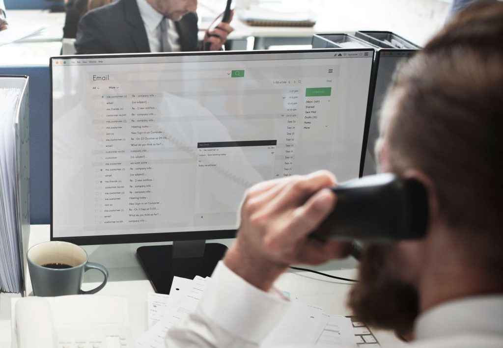 A close up of a person answering a phone in front of a large desktop screen.
