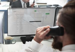 A close up of a person answering a phone in front of a large desktop screen.