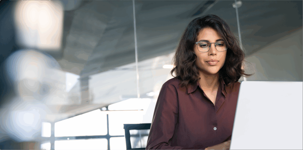 A woman wearing a burgundy shirt and glasses looking at a laptop screen in front of her.