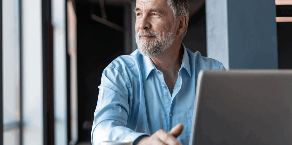 Older man sitting with his laptop and staring out a window.