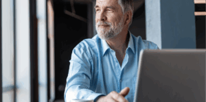 Older man sitting with his laptop and staring out a window.