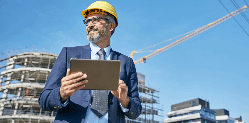 Man in a suit and a hard hat walking through a construction site with a laptop.