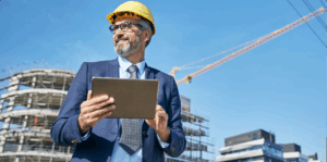 Man in a suit and a hard hat walking through a construction site with a laptop.