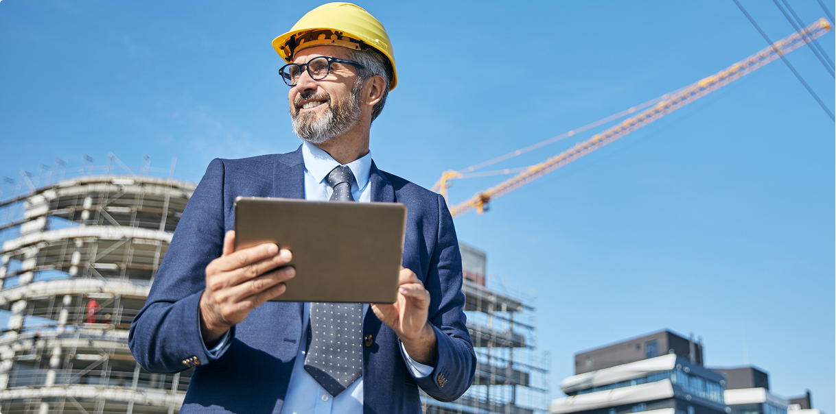 Man in a suit and a hard hat walking through a construction site with a laptop.