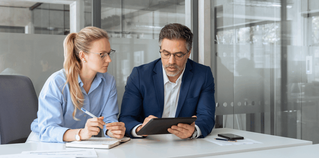 Two coworkers sitting at a boardroom table and looking at an iPad screen.