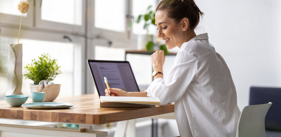 A woman at a desk writing something down, with a laptop open in front of her.