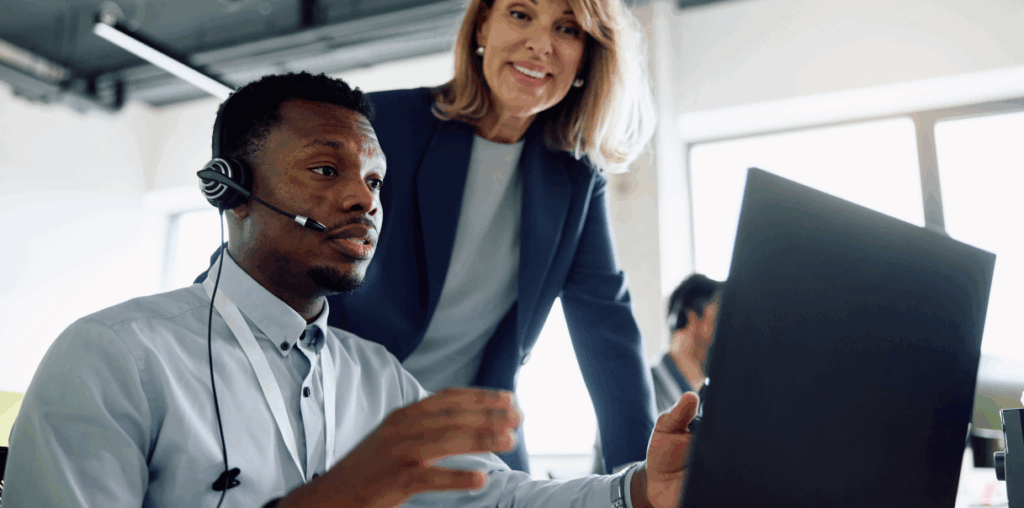 A person wearing a headset looking at a screen and a coworker smiling behind them.