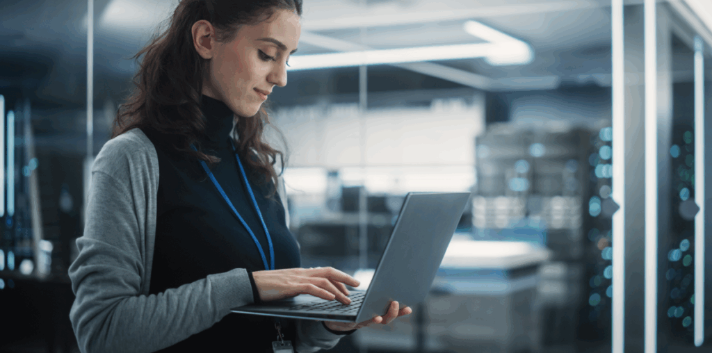 Woman standing up and holding a laptop in one hand while typing with the other.