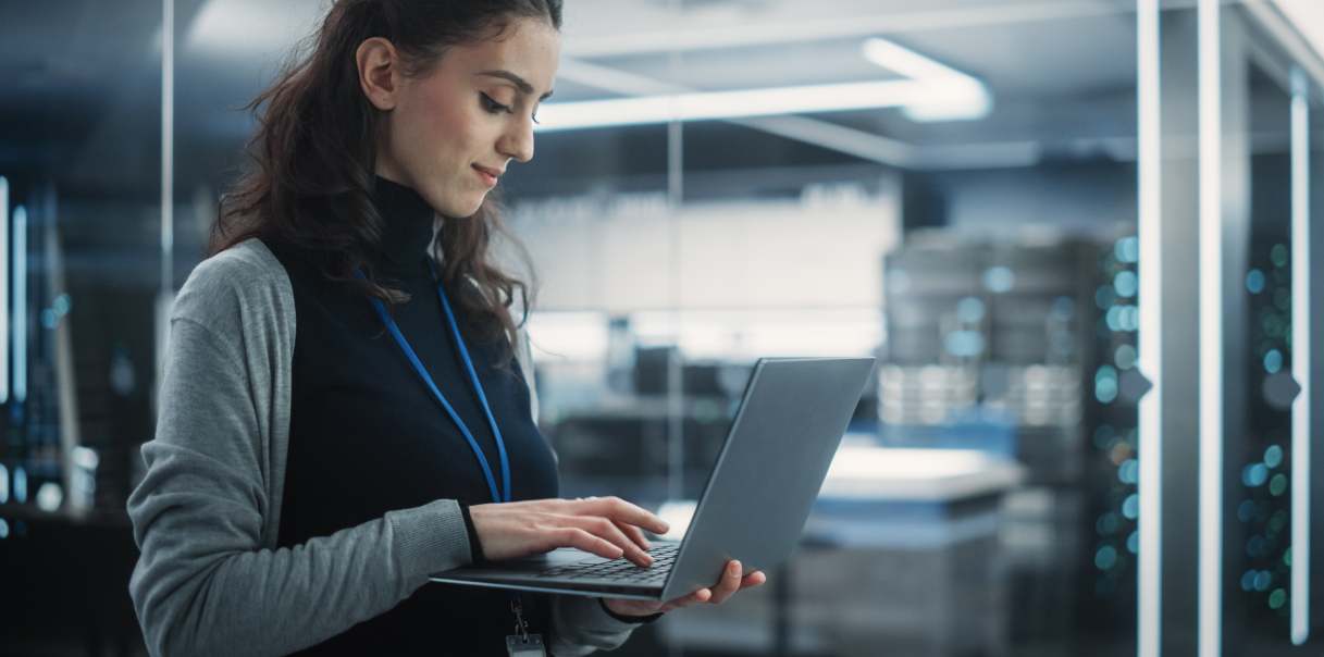 Woman standing up and holding a laptop in one hand while typing with the other.