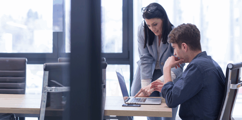 Two coworkers at a workspace. One is standing, the other is sitting, and both are looking at a laptop screen.