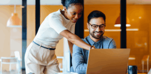 One employee sitting down while another stands next to them. Both are looking at a laptop screen.