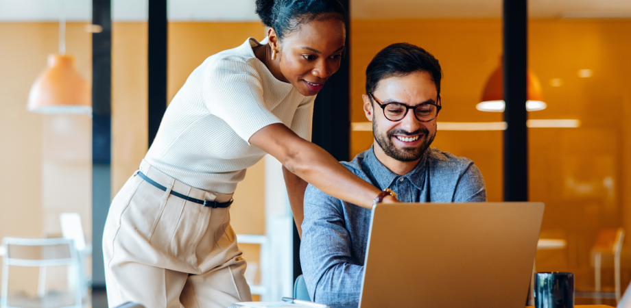 One employee sitting down while another stands next to them. Both are looking at a laptop screen.