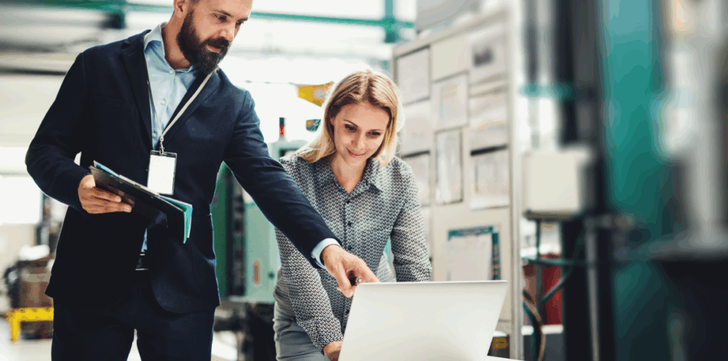 An employee using a laptop while another employee carrying a tablet stands next to them and points at the laptop screen.