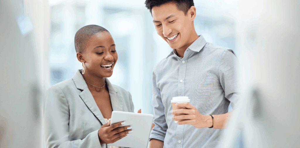 Two coworkers standing and smiling at a tech device.