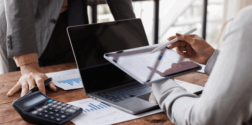 Person holding an iPad in front of a laptop screen. Opposite them is a person with their hands resting on a desk.