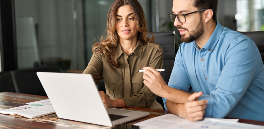 A man and woman are in discussion at a table with an open laptop.