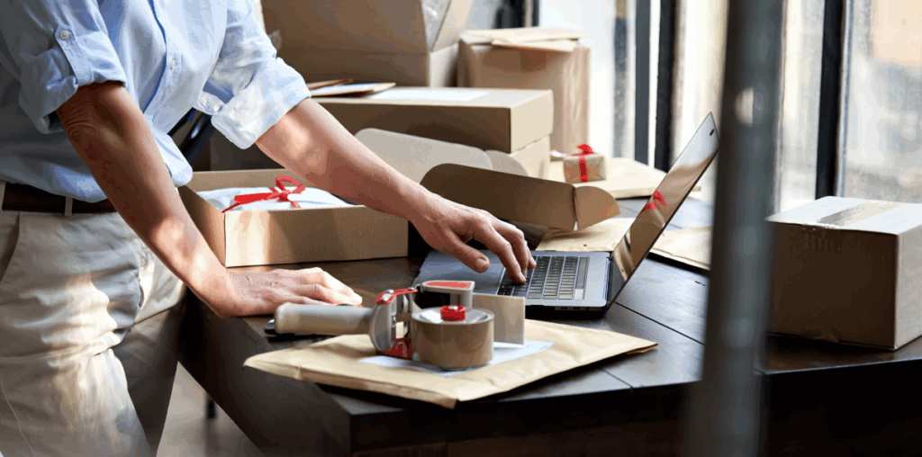 Person working on a laptop, standing next to cardboard boxes and packing tape.
