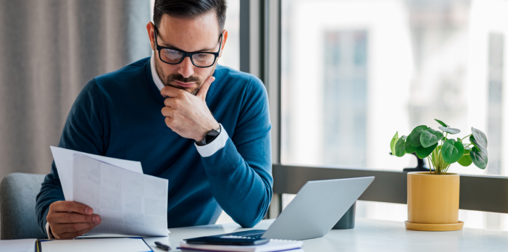 A man deep in thought, holding papers and sitting next to a laptop