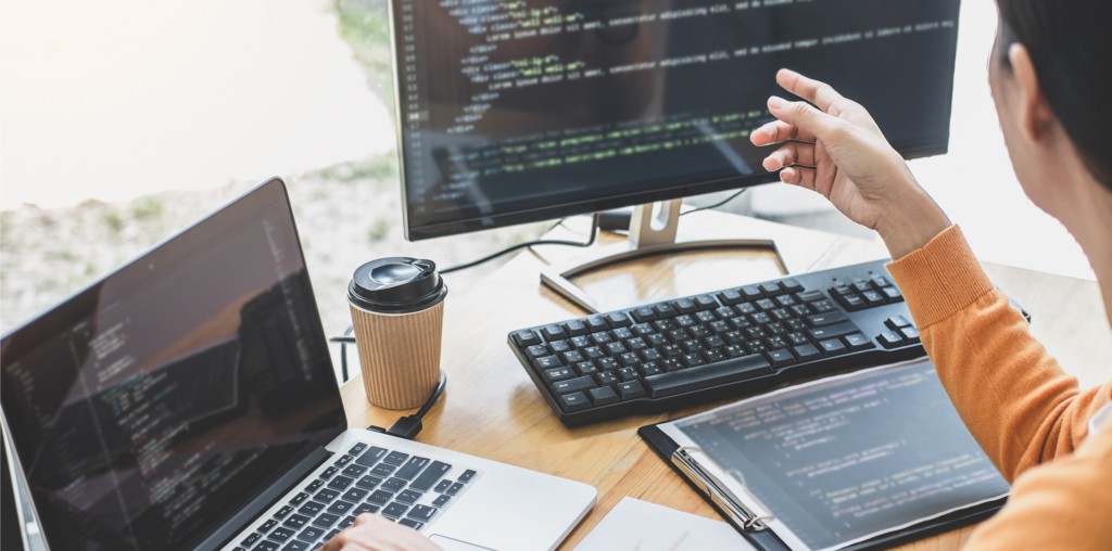 A programmer points at a computer monitor displaying code while working at a desk with a laptop, a keyboard, and a coffee cup.
